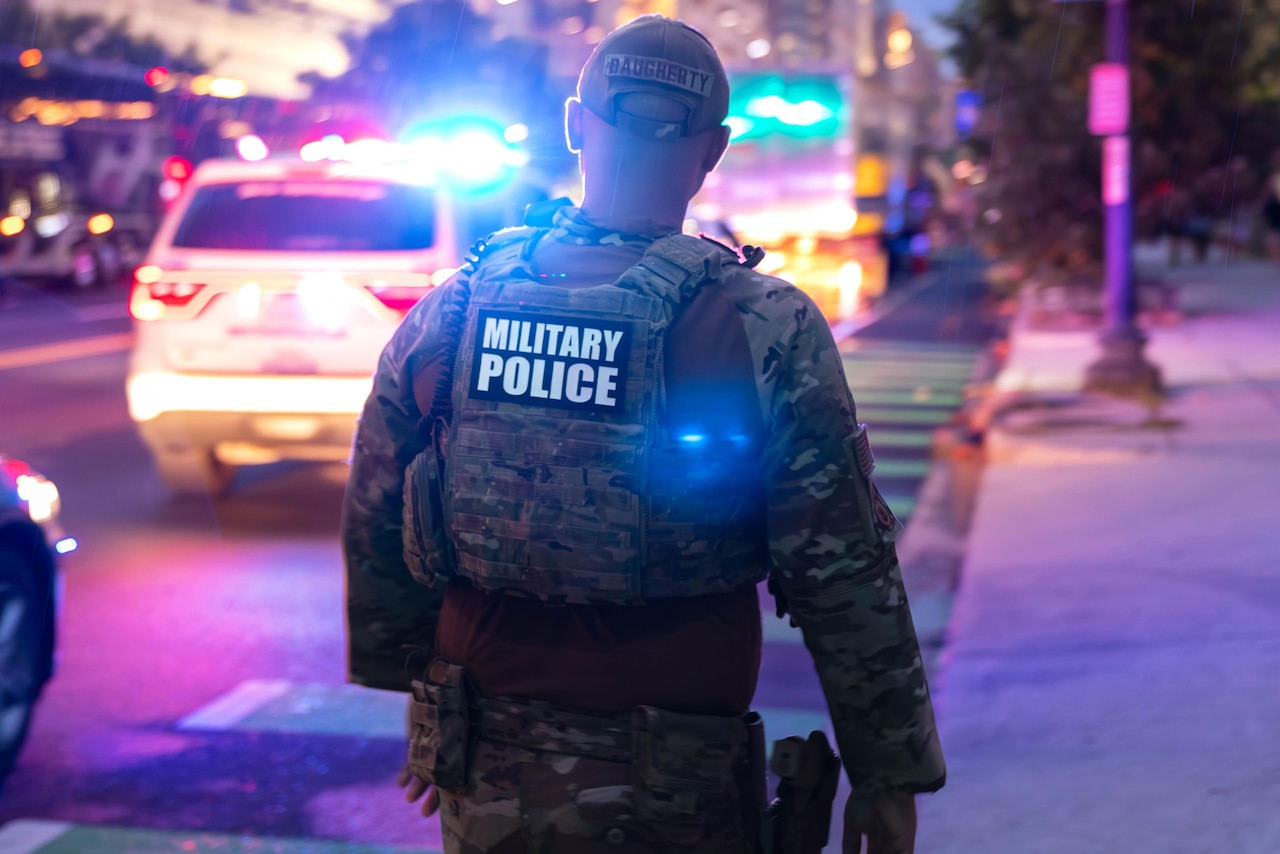 An airman in a camouflage military uniform walks down a sidewalk in a city. There is a police vehicle with its lights flashing in the background.