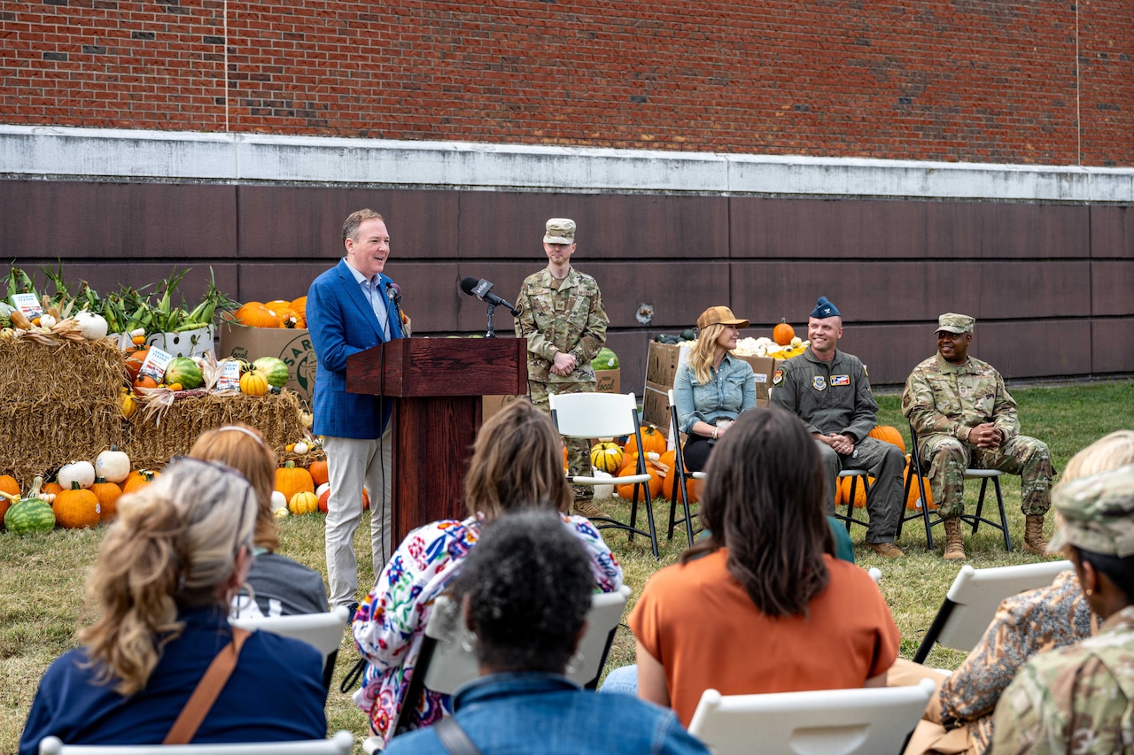 A man in business attire stands at a lectern and speaks to a crowd. There is a person in a camouflage military uniform standing to the right of the lectern and two people in military uniforms and one in casual attire sitting to the right of the lectern. There are pumpkins and other produce displayed in the background.