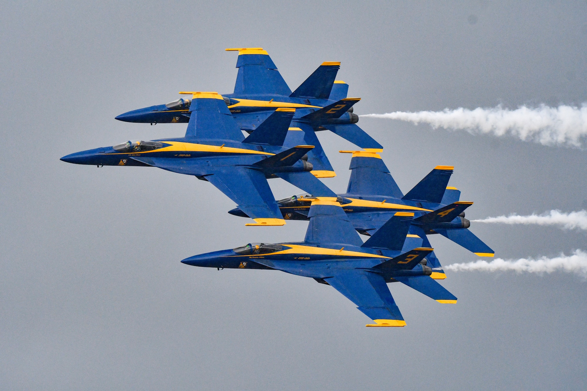 Four Blue Angels flying in a cloudy-gray sky, white smoke trailing behind.
