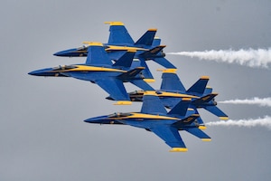 Four Blue Angels flying in a cloudy-gray sky, white smoke trailing behind.