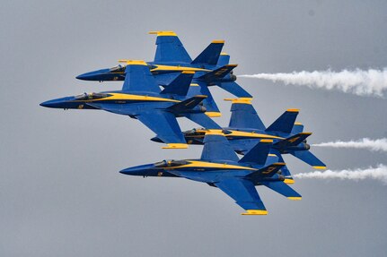 Four Blue Angels flying in a cloudy-gray sky, white smoke trailing behind.