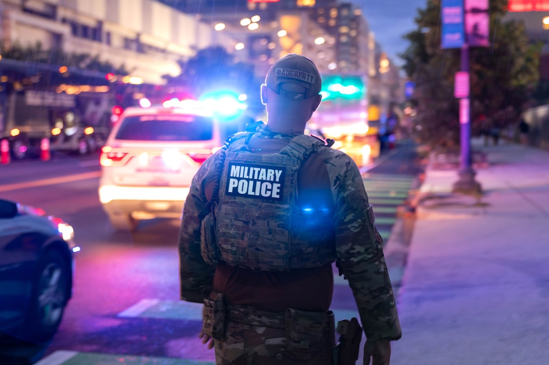 A guardsman wearing a vest that says "MILITARY POLICE" walks on a sidewalk next to cars bathed in blue and red light, as seen from behind.