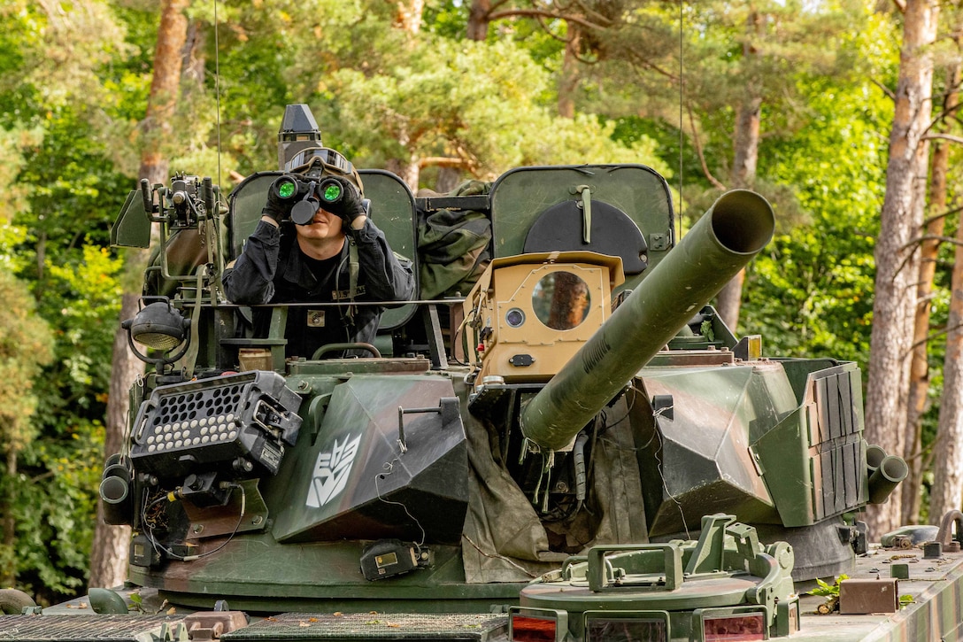 A soldier sitting in a green tank uses binoculars while in front of a forest setting during the day, as seen from head on.