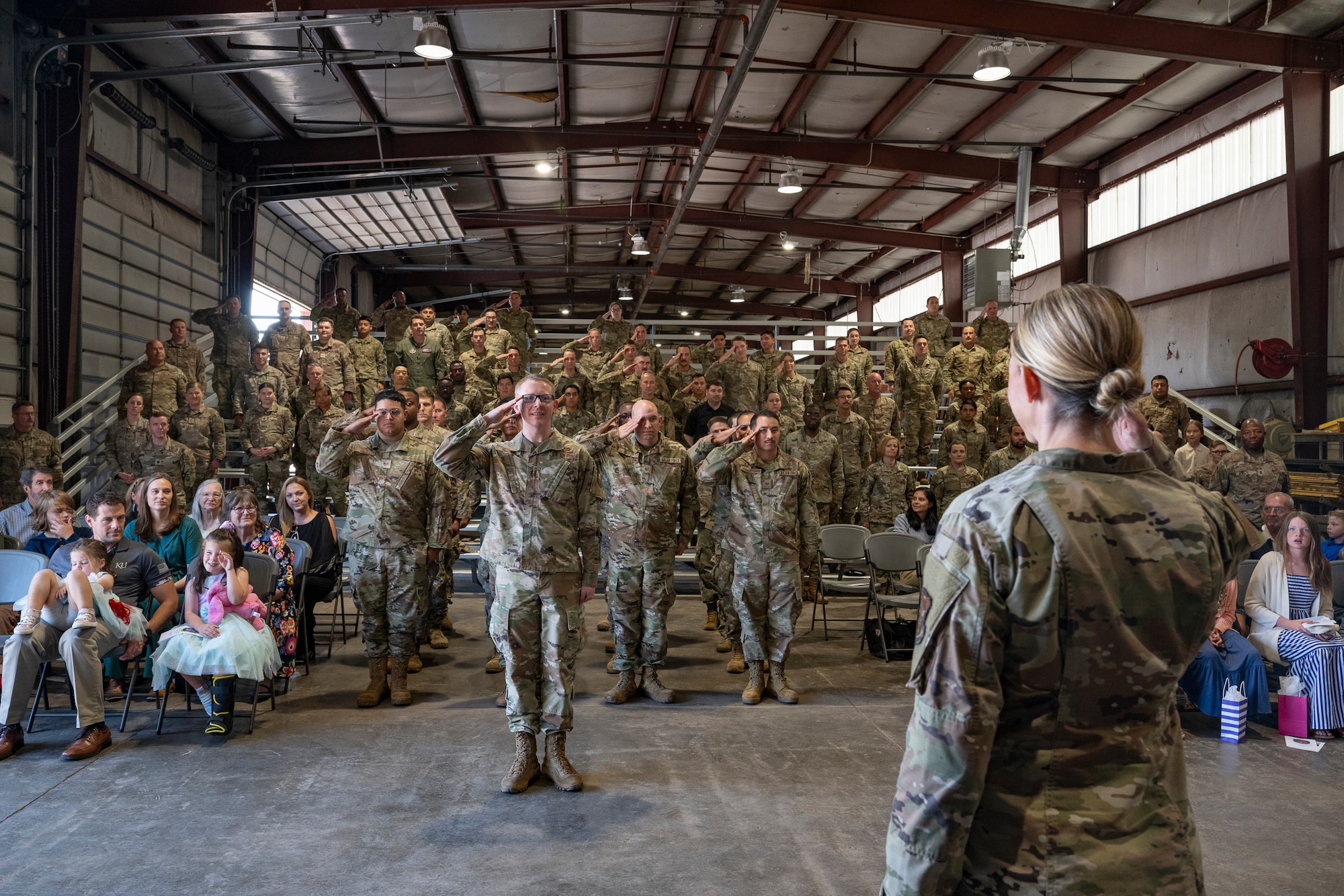 A crowd in a warehouse with military servicemembers in attendance saluting a woman at the front of the room.