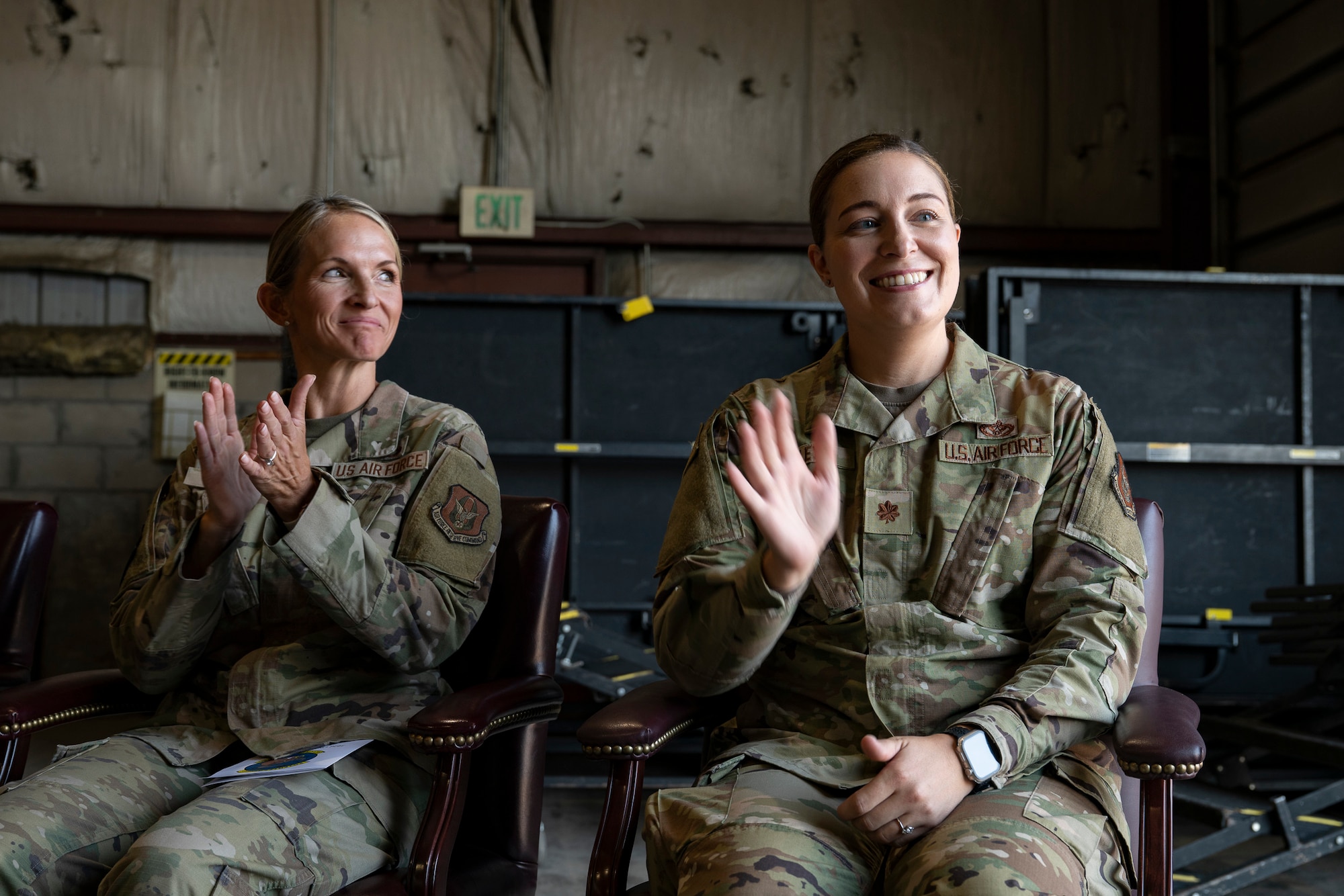 Two servicemembers in uniform seated in chairs, one waving and the other clapping.