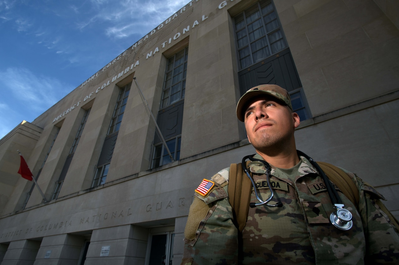 A person wearing a camouflage military uniform stands outside in front of a building and gazes up into the distance.
