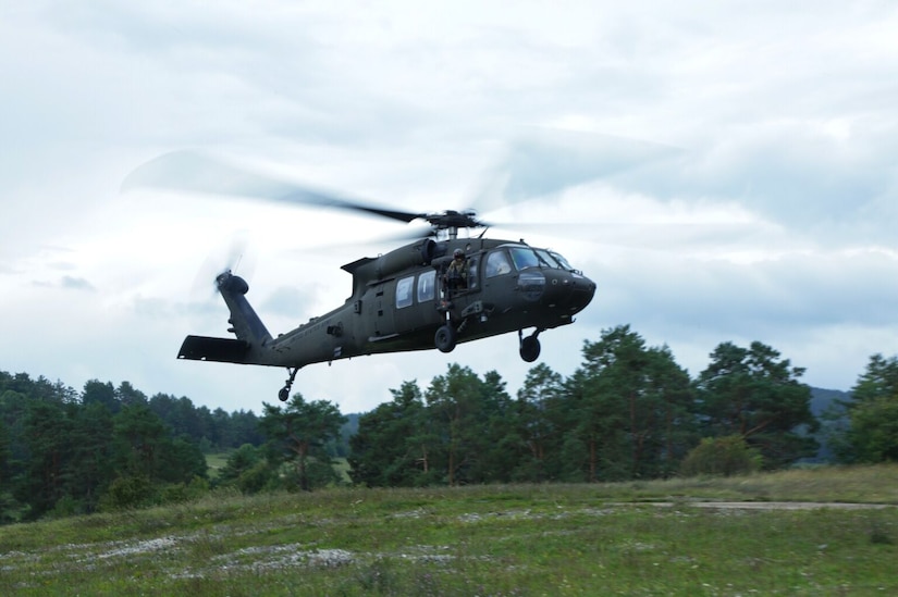 A soldier wearing a camouflage military uniform leans out the window of a helicopter that is hovering above the ground. There are trees in the background and an open field in the foreground.