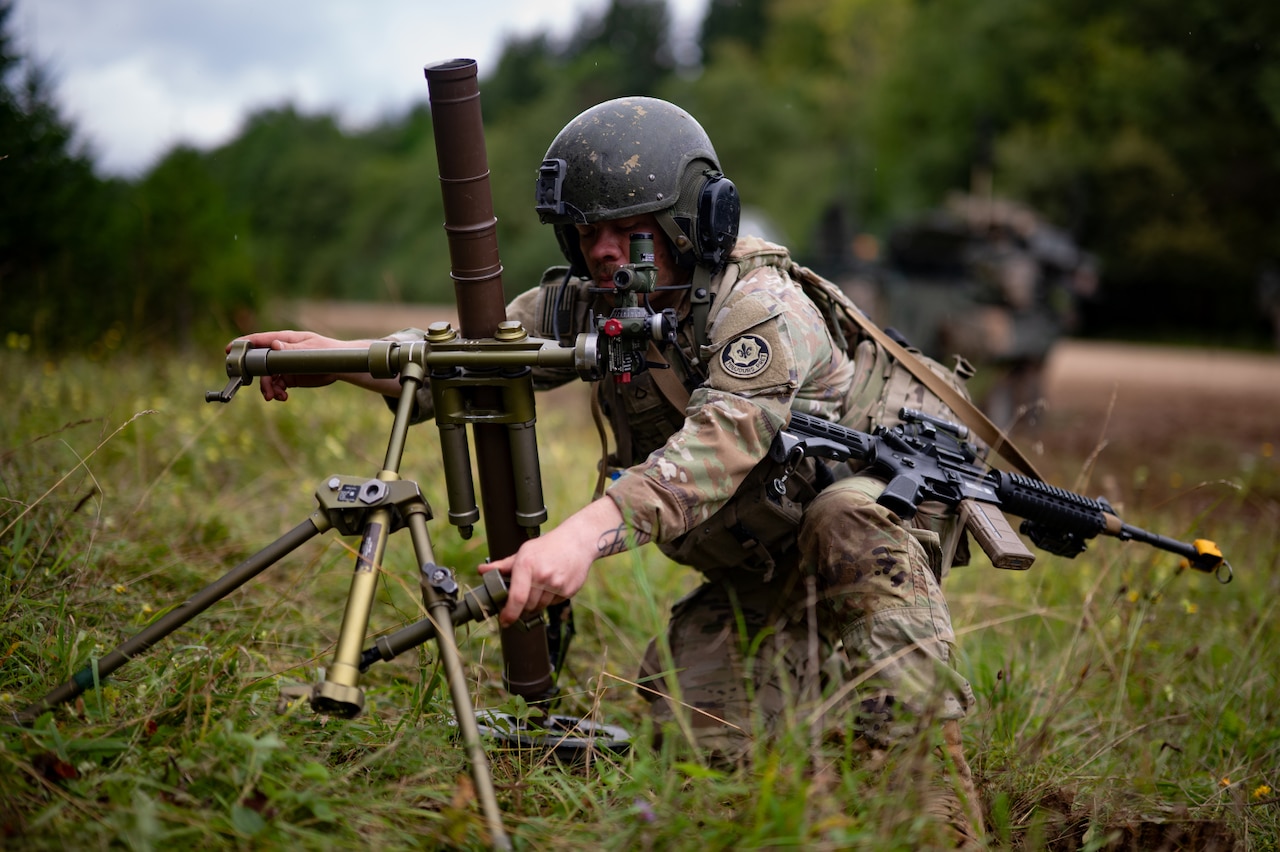 A soldier wearing a camouflage military uniform and a helmet places a mortar firing device on the ground in a grassy area. The soldier is carrying a machine gun on his shoulder and a backpack.