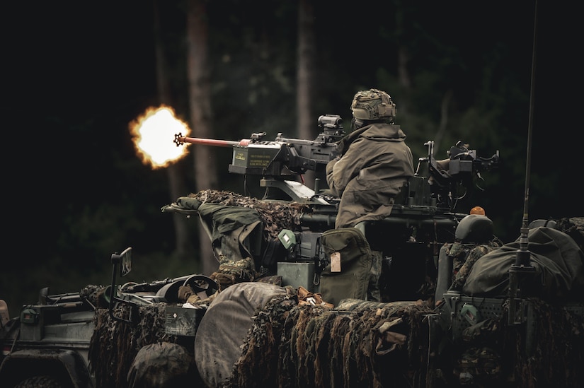 A soldier wearing a camouflage military uniform fires a large gun from the top of a military vehicle.