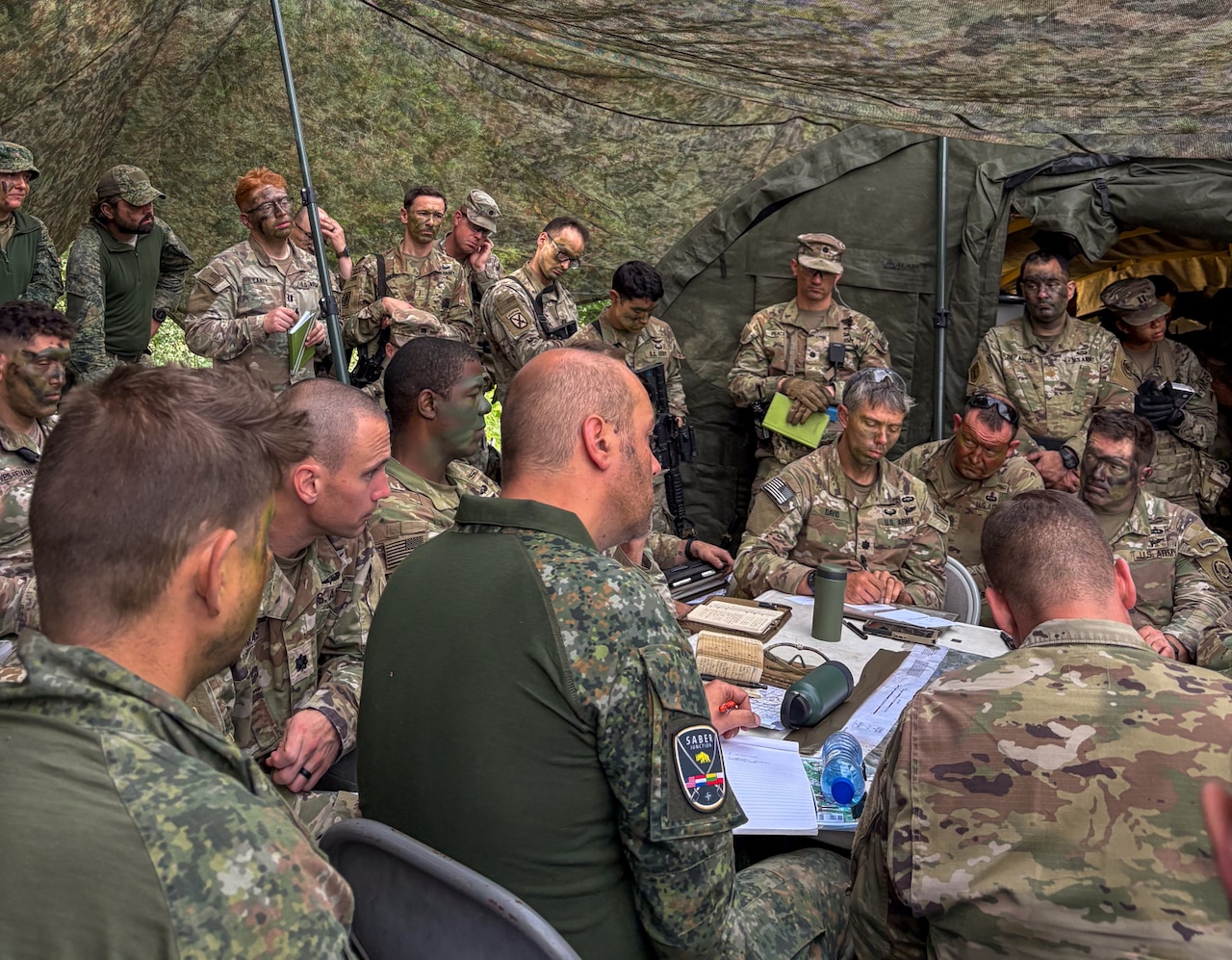 Several soldiers in camouflage military uniforms stand in a tent as more soldiers in similar attire sit at a table. Some of the soldiers have camouflage paint of their faces.