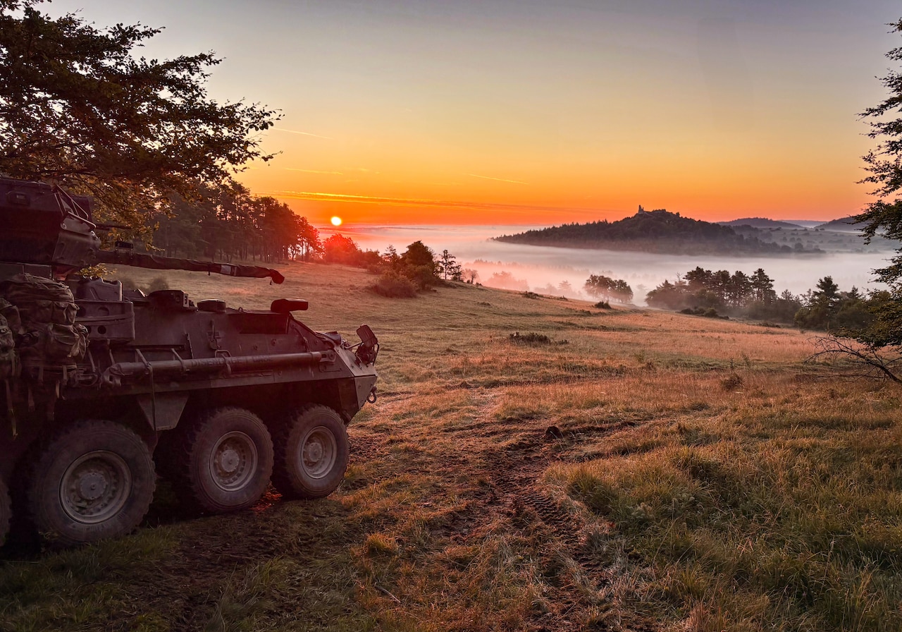 A tank parked on a hill overlooks a fog-covered valley at sunrise.