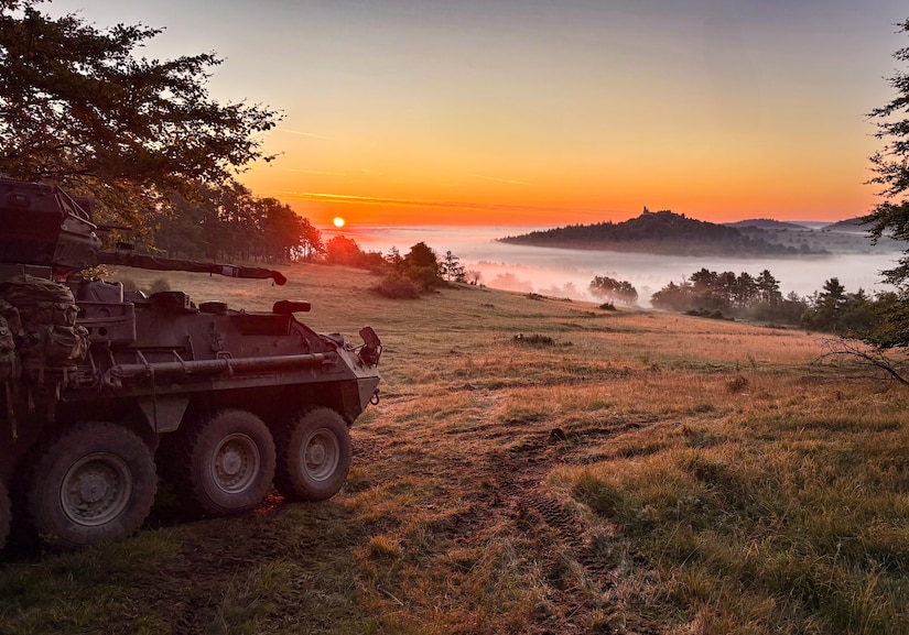 A tank parked on a hill overlooks a fog-covered valley at sunrise.