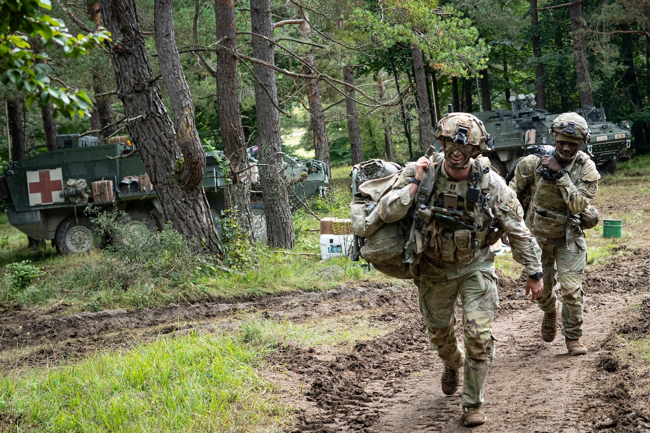Two soldiers carrying packs walk in a forested area away from a military ambulance parked in the background.