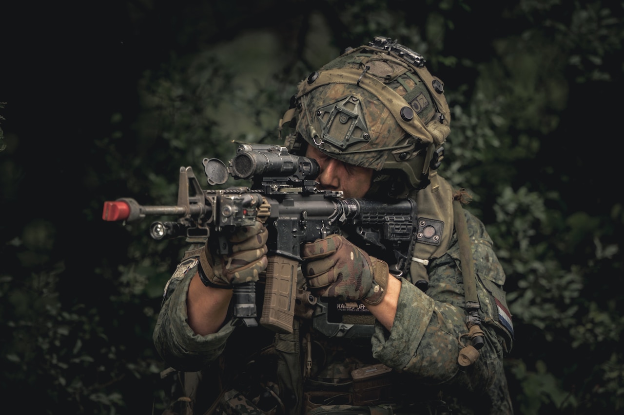 A soldier wearing a camouflage military uniform and a helmet holds a machine gun up to his eye as he aims the weapon.