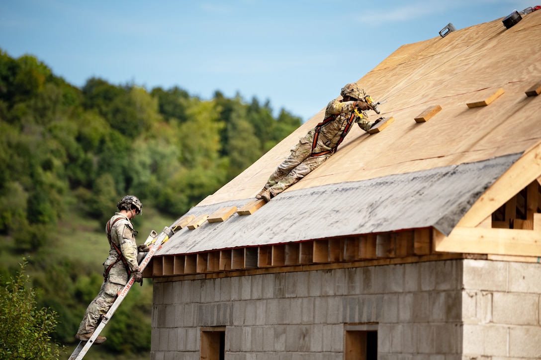 A soldier on a ladder watches as a fellow soldier uses a drill on a wooden roof.