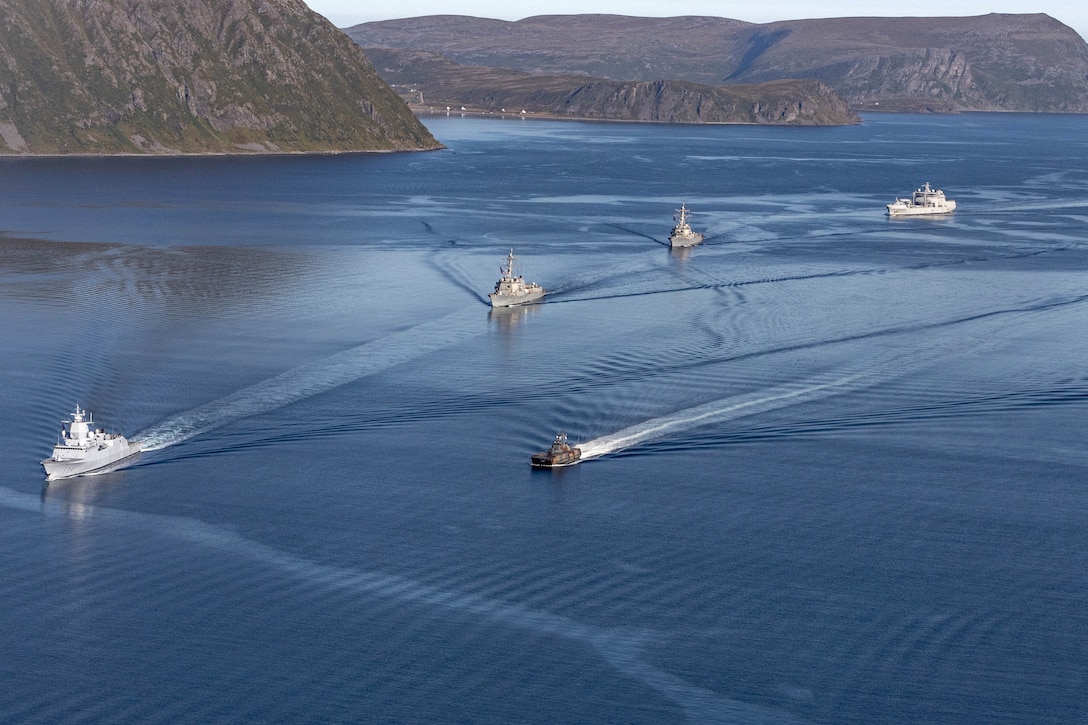 Five ships sail in formation in blue waters near with the coastline in the background.