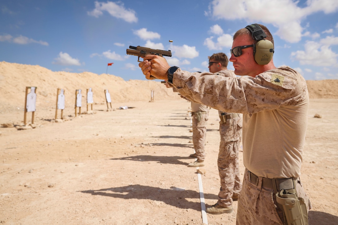 Ammo flies in the air as a Marine fires a weapon in a desert-like area with fellow Marines under a blue sky.