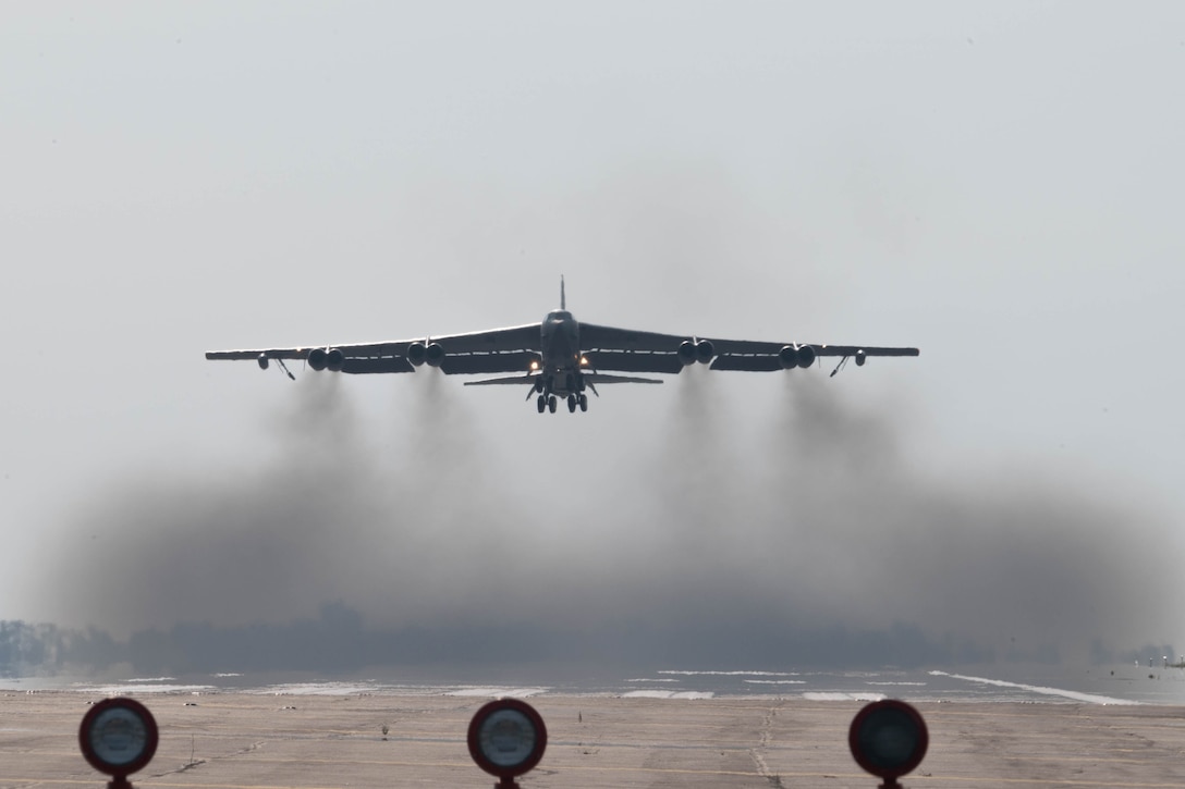 An aircraft takes off from a runway, leaving grey smoke behind on an overcast day, as seen from head on.