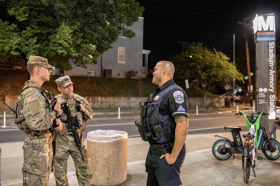 Two guardsmen speak with a police officer while all three stand on a sidewalk next to a road, with two bikes and a Metro station sign to the right.