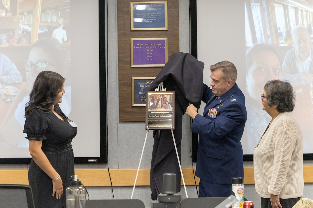 A man, wearing a blue military uniform, unveils a plaque on a stand as two women stand near him