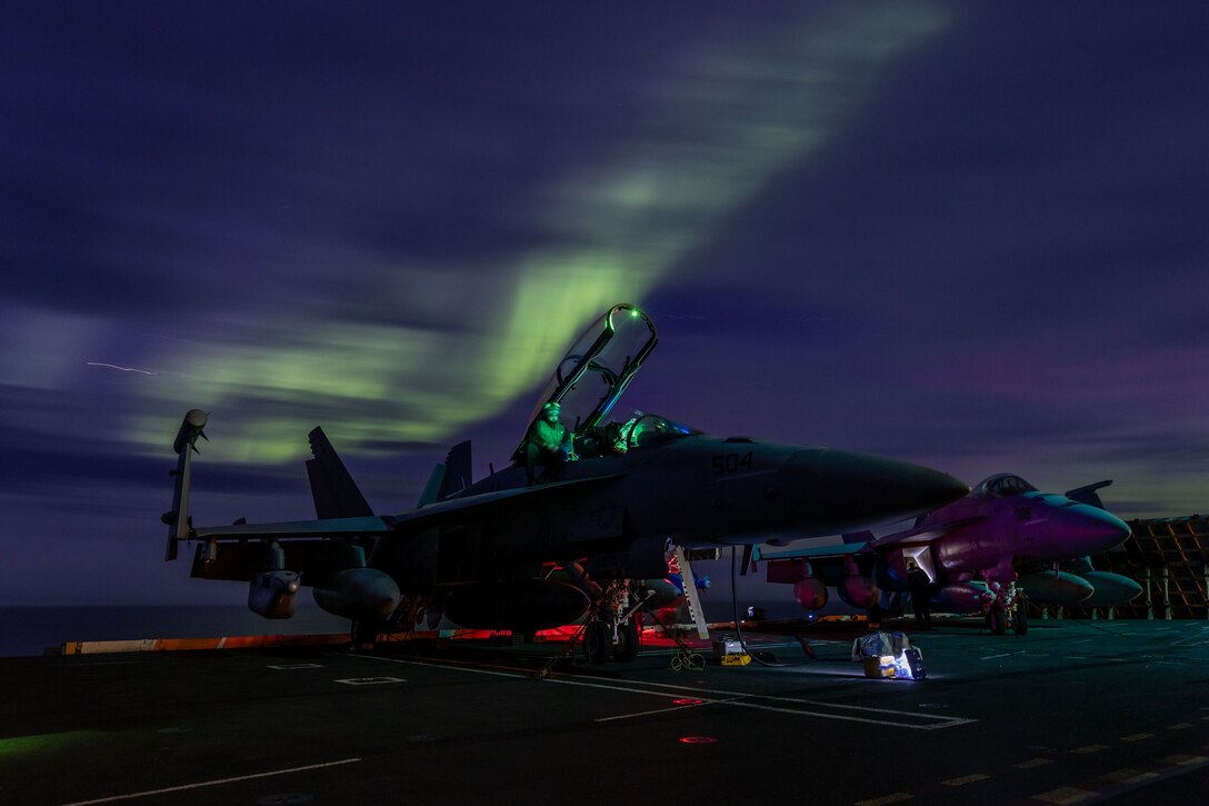 Northern Lights in an otherwise dark sky illuminate a ship's deck where sailors work on an aircraft.