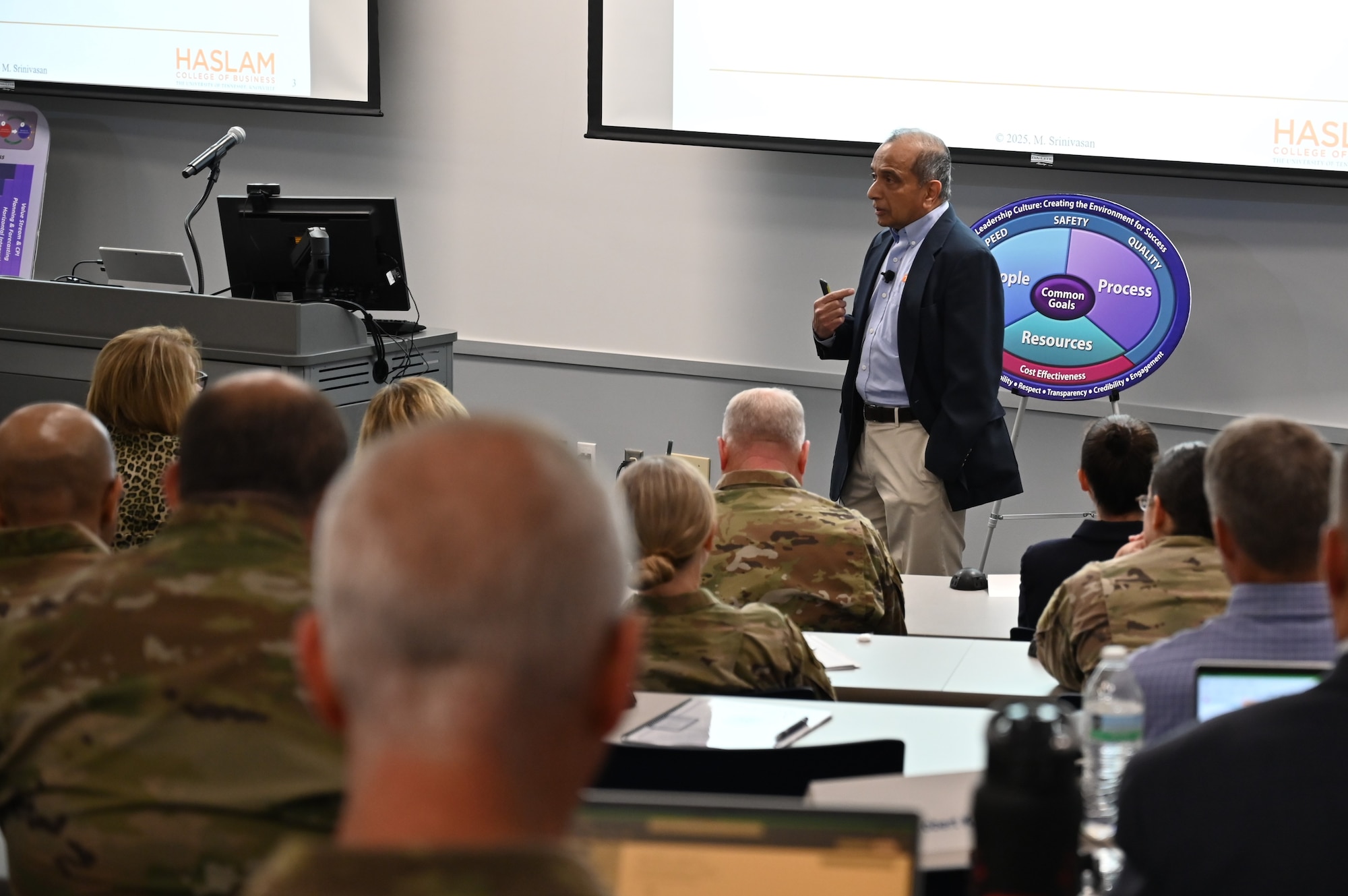 A civilian presenter speaks to civilian and uniformed civilian personnel.