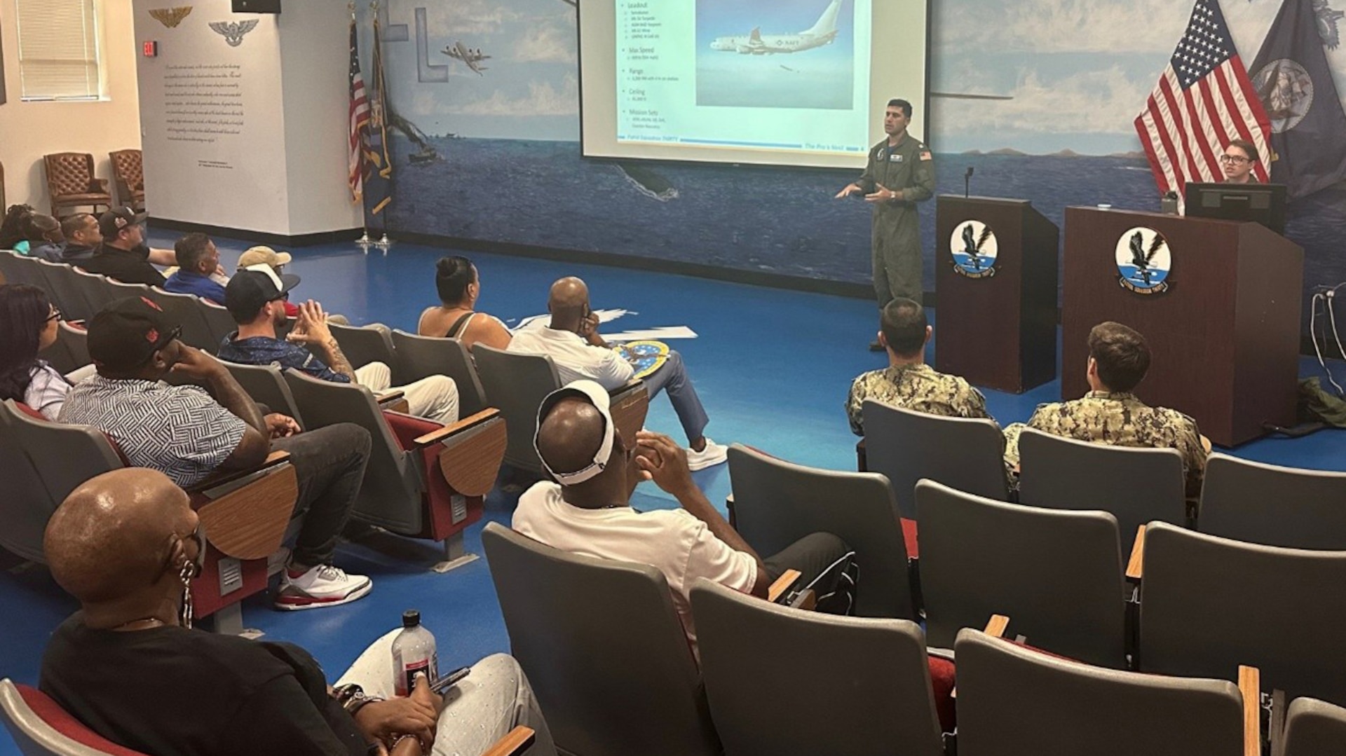 A group of employees listen to a sailor in an auditorium.