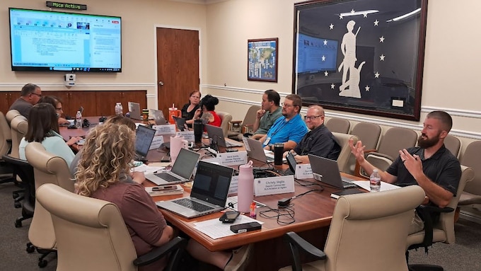 A group of employees sit around a conference table discussing content on a powerpoint slide.