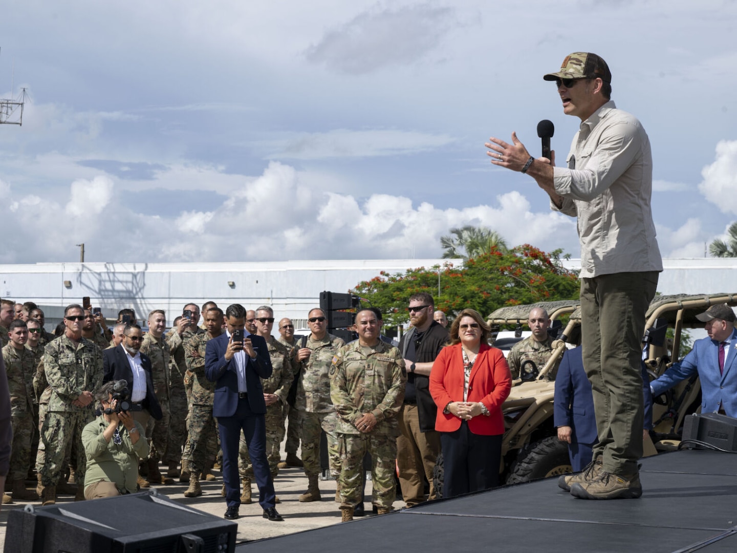 A man in civilian attire, including a baseball cap, is standing on a stage speaking into a microphone while addressing a group of people with their backs to the camera. Behind him is a large logo on the wall that reads "USS Iwo Jima."