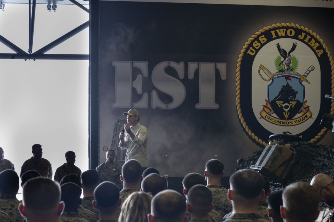 A man in civilian attire, including a baseball cap and sunglasses, is standing on a stage outside speaking into a microphone while addressing a group of roughly two dozen people. Many people are wearing camouflage military uniforms, while others are in civilian attire.