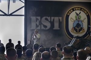 A man in civilian attire, including a baseball cap and sunglasses, is standing on a stage outside speaking into a microphone while addressing a group of roughly two dozen people. Many people are wearing camouflage military uniforms, while others are in civilian attire.