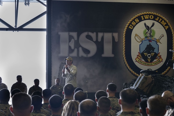 A man in civilian attire, including a baseball cap and sunglasses, is standing on a stage outside speaking into a microphone while addressing a group of roughly two dozen people. Many people are wearing camouflage military uniforms, while others are in civilian attire.