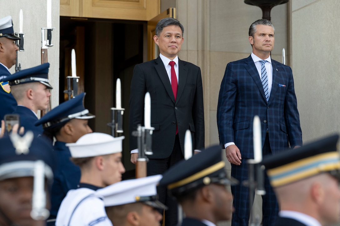 Secretary of War Pete Hegseth and another leader stand outside an open door with troops on steps flanking their right side.