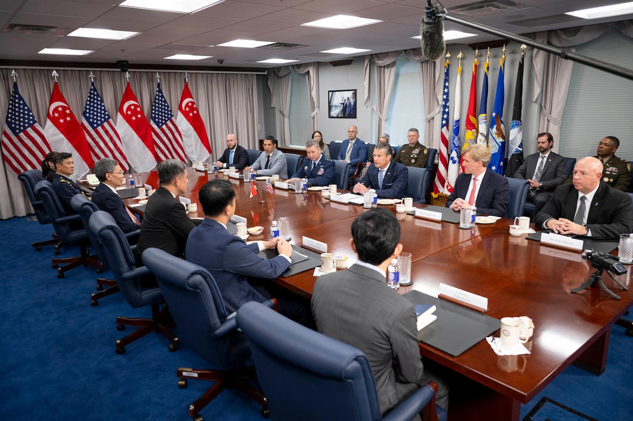 Several people in business suits and two in military dress uniforms sit around a large table with flags in the background.