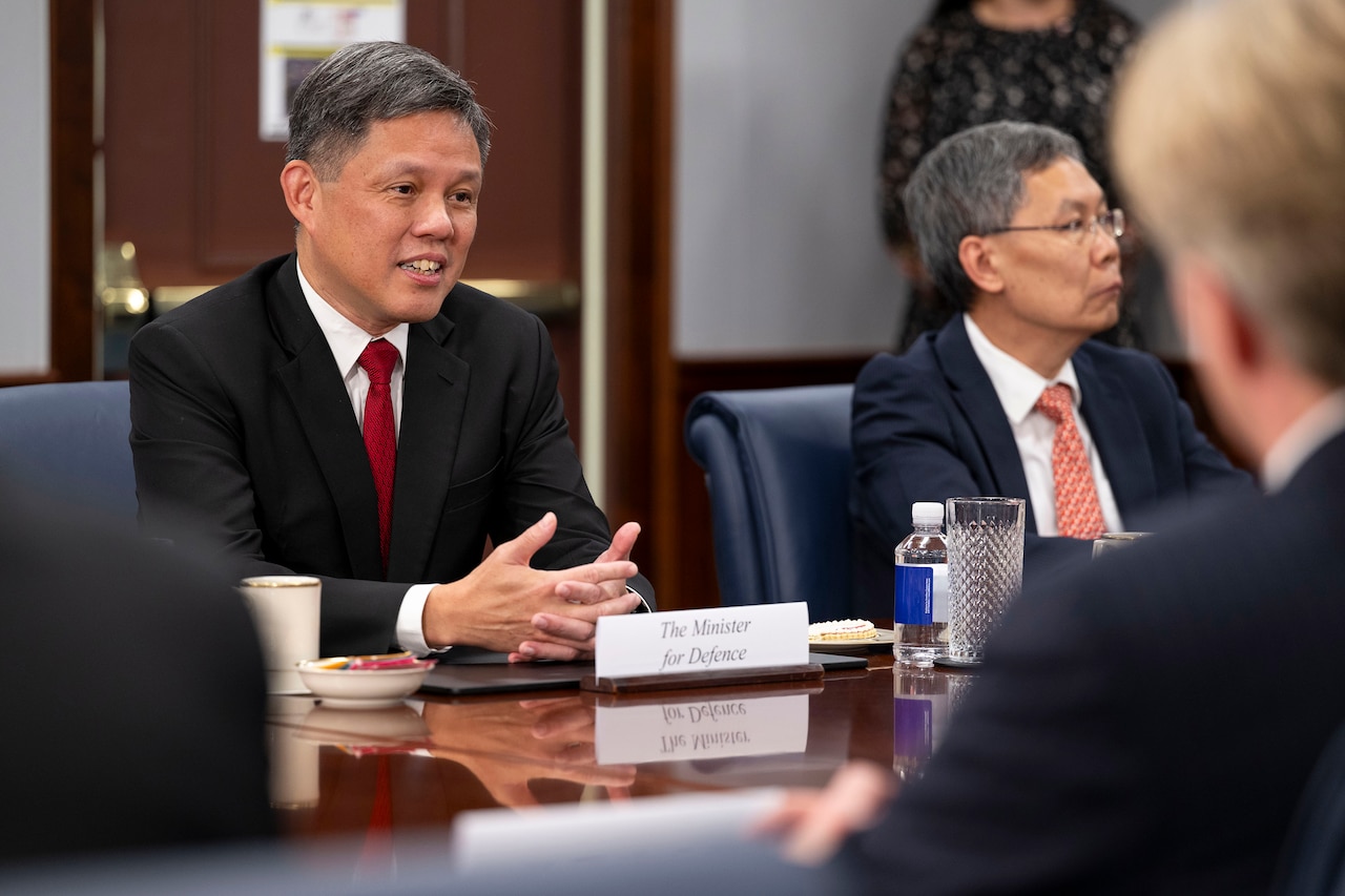 A civilian in a business suit speaks while seated at a table with other officials.
