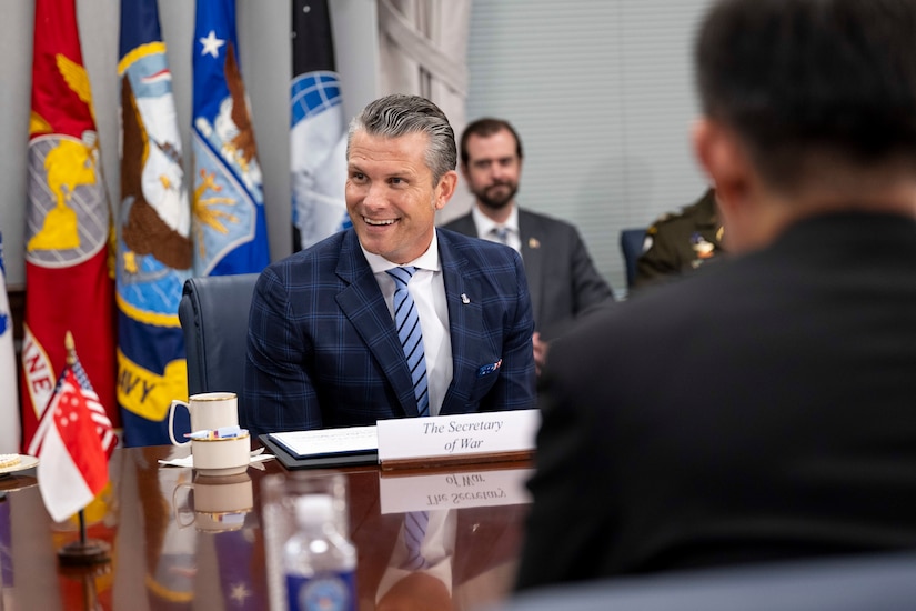 Secretary of War Pete Hegseth smiles while sitting at a table with other officials and flags in the background.