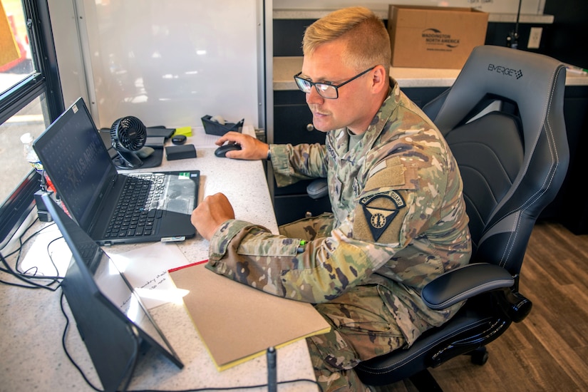 A soldier in a camouflage military uniform sits at a desk with a computer and a tablet in front of him.