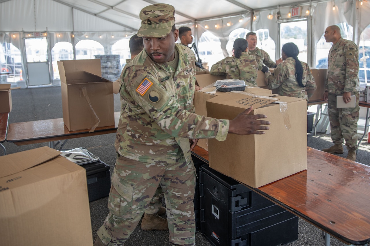 A soldier in a camouflage military uniform reaches for a box that is on a nearby table. There are several soldiers in similar attire standing around a table full of boxes in the background.