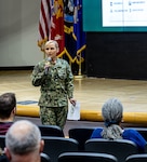 Woman with blonde hair in military uniform stands in front of stage speaking to audience, holding microphone with one hand and eyeglasses and papers in the other.