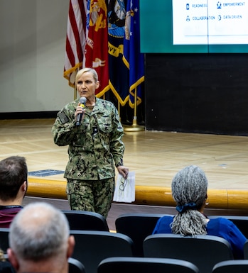 Woman with blonde hair in military uniform stands in front of stage speaking to audience, holding microphone with one hand and eyeglasses and papers in the other.