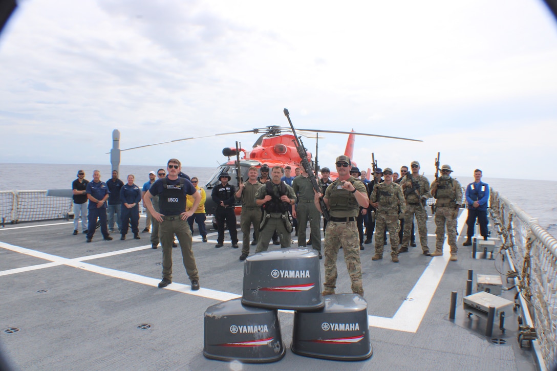 Coast Guard crews from the Coast Guard Helicopter Interdiction Tactical Squadron, Coast Guard Tactical Law Enforcement Team – South, Coast Guard Cutter Midgett (WMSL 757) helicopter tie-down members and unmanned aerial vehicle personnel pose for a group photo aboard Midgett from behind three bullet-damaged outboard engine cowlings while underway in the Eastern Pacific Ocean, Aug. 28, 2025. On Aug. 25, HITRON used airborne use of force to stop the non-compliant vessel, marking the unit’s 1,000th drug interdiction since the unit’s inception in 1999, which resulted in Midgett crew members seizing approximately 3,606 pounds of suspected cocaine worth an estimated $46 million and apprehending six suspected narco-traffickers. (U.S. Coast Guard photo)