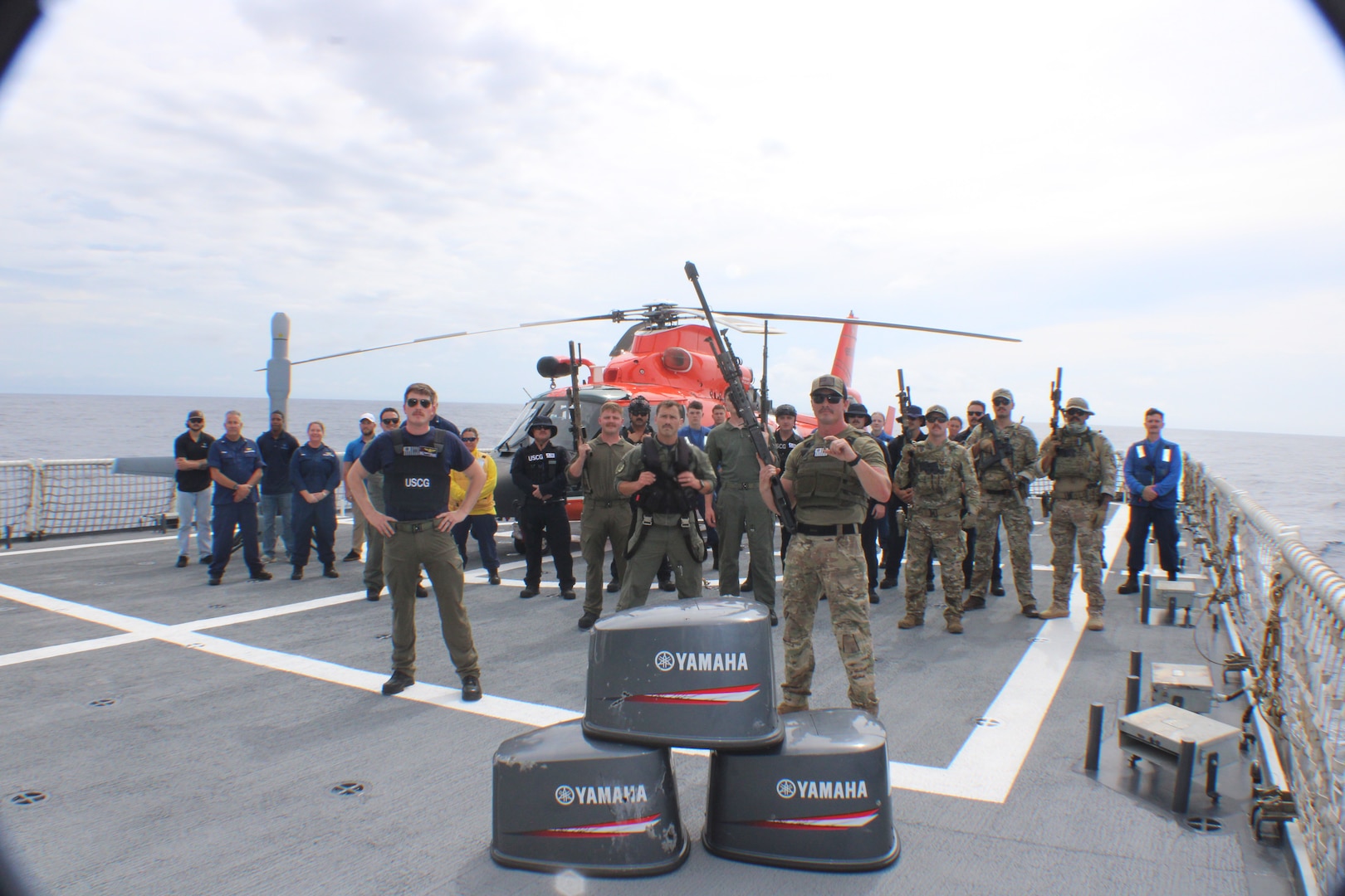 Coast Guard crews from the Coast Guard Helicopter Interdiction Tactical Squadron, Coast Guard Tactical Law Enforcement Team – South, Coast Guard Cutter Midgett (WMSL 757) helicopter tie-down members and unmanned aerial vehicle personnel pose for a group photo aboard Midgett from behind three bullet-damaged outboard engine cowlings while underway in the Eastern Pacific Ocean, Aug. 28, 2025. On Aug. 25, HITRON used airborne use of force to stop the non-compliant vessel, marking the unit’s 1,000th drug interdiction since the unit’s inception in 1999, which resulted in Midgett crew members seizing approximately 3,606 pounds of suspected cocaine worth an estimated $46 million and apprehending six suspected narco-traffickers. (U.S. Coast Guard photo)