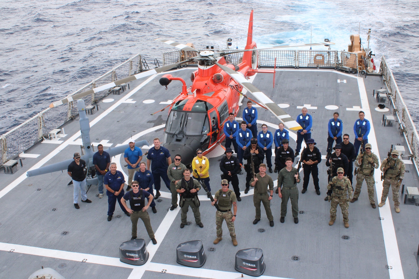 Coast Guard crews from the Coast Guard Helicopter Interdiction Tactical Squadron, Coast Guard Tactical Law Enforcement Team – South, Coast Guard Cutter Midgett (WMSL 757) helicopter tie-down members and unmanned aerial vehicle personnel pose for a group photo aboard Midgett from behind three bullet-damaged outboard engine cowlings while underway in the Eastern Pacific Ocean, Aug. 28, 2025. On Aug. 25, HITRON used airborne use of force to stop the non-compliant vessel, marking the unit’s 1,000th drug interdiction since the unit’s inception in 1999, which resulted in Midgett crew members seizing approximately 3,606 pounds of suspected cocaine worth an estimated $46 million and apprehending six suspected narco-traffickers. (U.S. Coast Guard photo)
