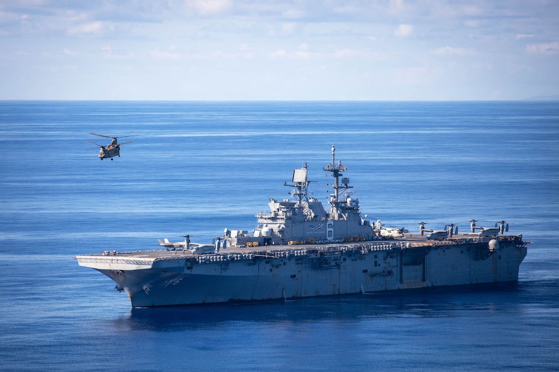 A helicopter flies over a ship transiting a body of water during the day.