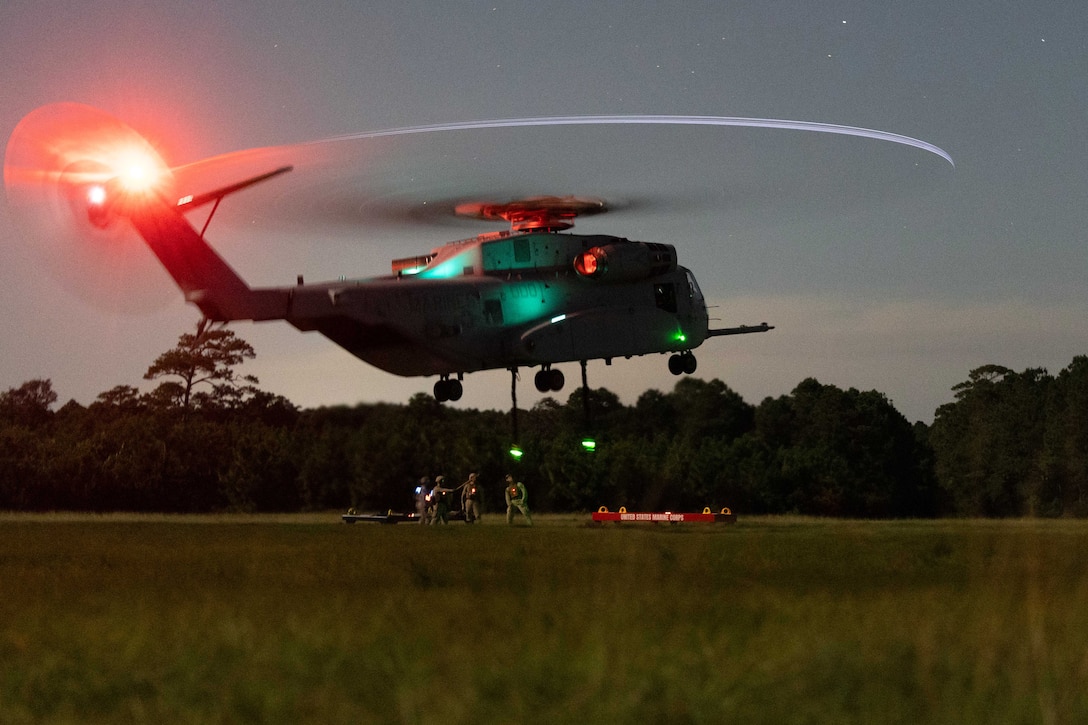 Marines in a dark field prepare to load a hovering helicopter illuminated by green and red lights.