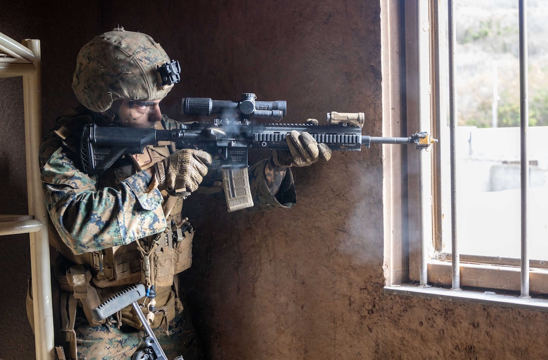 A Marine in tactical gear aims a service weapon through an open window while standing inside a brown room during the day.