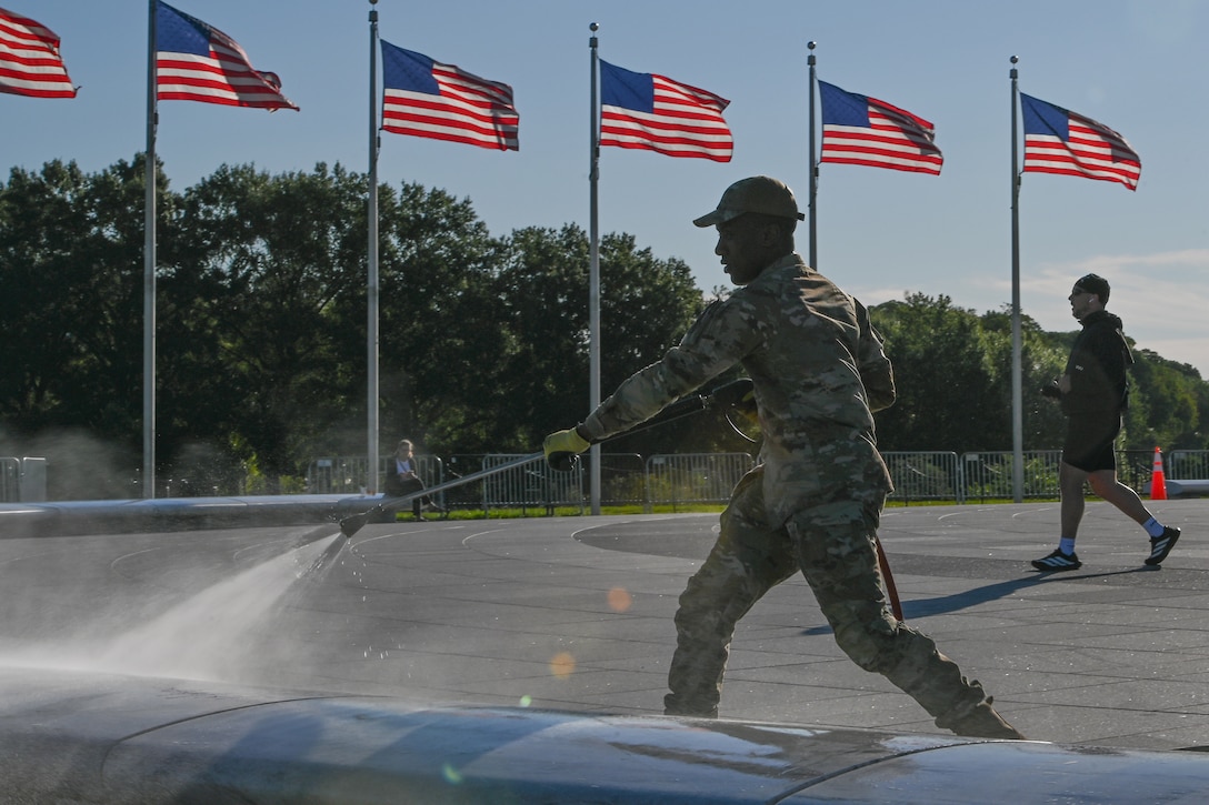 A guardsman uses a power hose to rinse off marble benches outside, as a person walks behind, with flag posts flying American flags in the background.