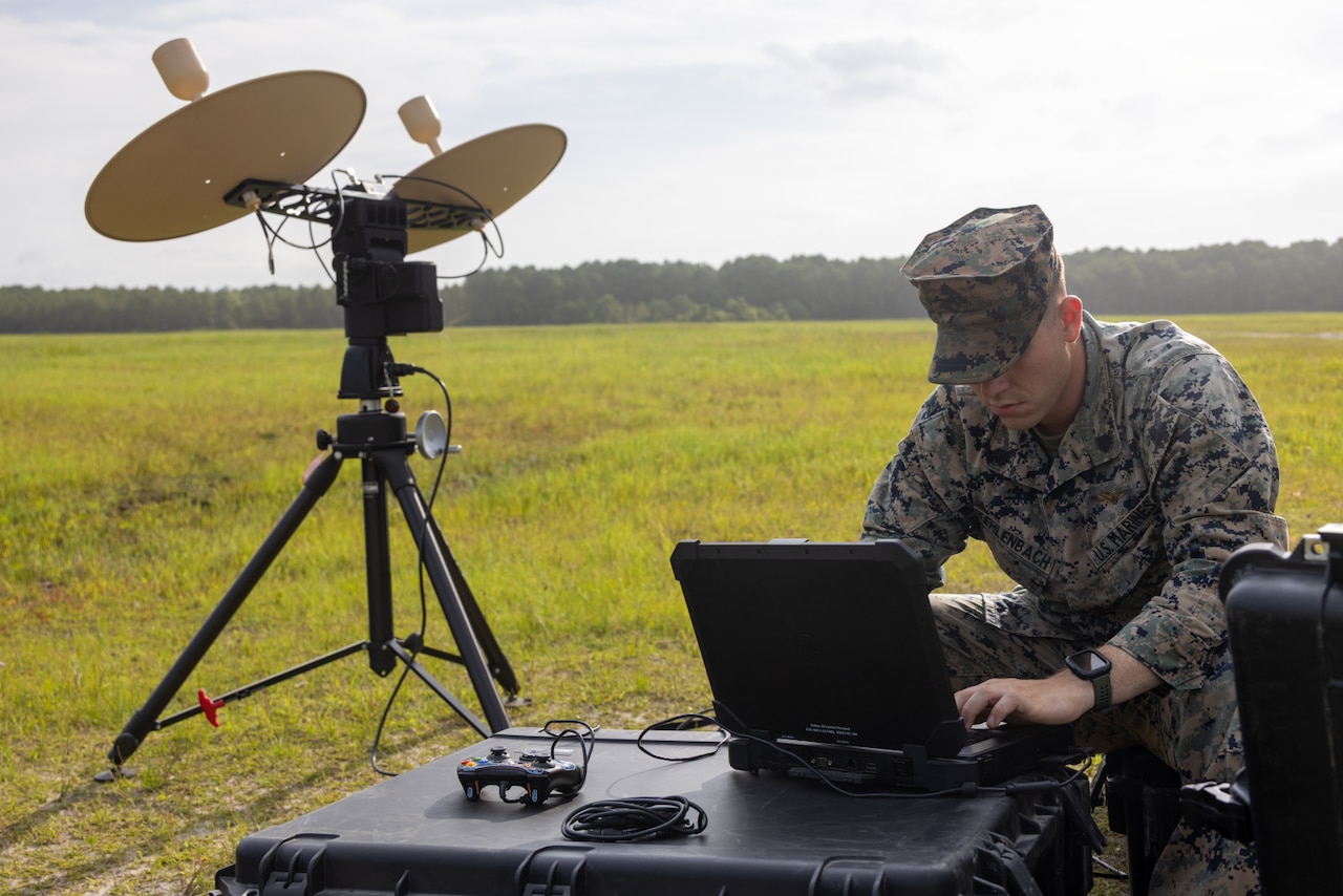A Marine wearing a camouflage military uniform sits on an equipment case while working on a computer that is on a case in front of him. There is unmanned aircraft system equipment to the left.