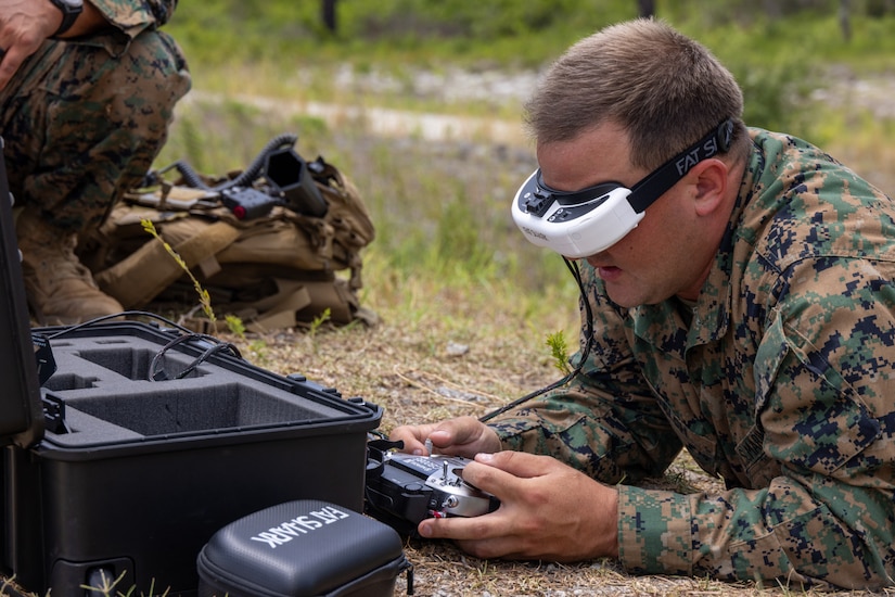 A Marine wearing a camouflage military uniform and goggles lies on the ground while using a remote for a drone. There is an equipment case in front of him.