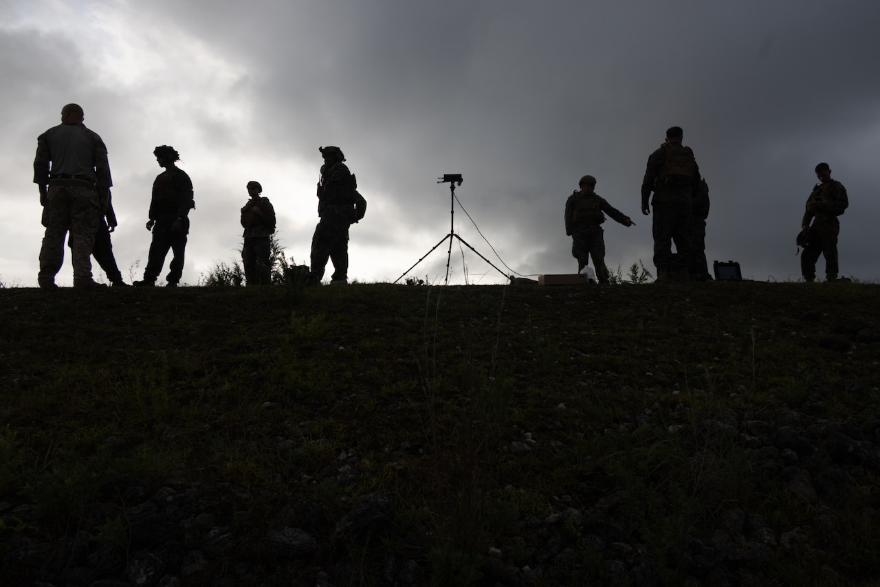A silhouette of people standing on top of a hill with equipment around them is shown.